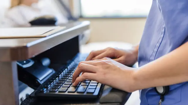 A healthcare worker in blye scrubs typing on a keyboard