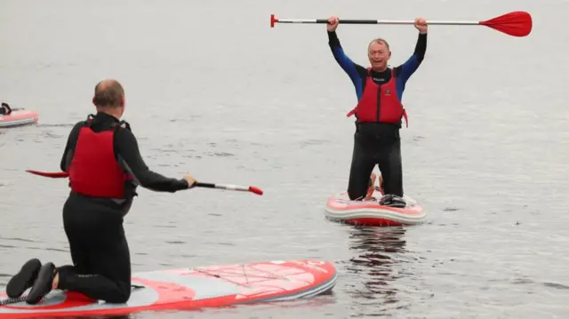 Tim Farron holds paddle in the air during paddleboarding in Windermere