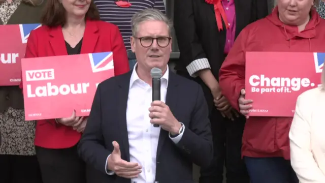 Labour leader Keir Starmer holding a microphone with a crowd of Labour party supporters holding red sign standing behind him