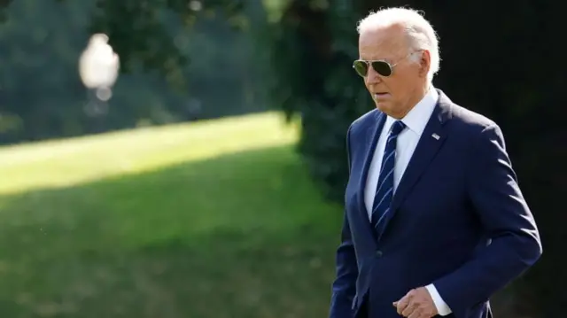 Joe Biden walking in front of some grass. His head is facing the floor and he is wearing sunglasses