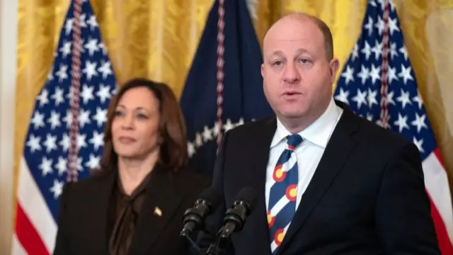 Polis speaks at a lectern, with Harris standing behind him