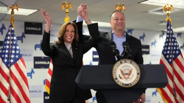 Kamala Harris on stage holding her hands in the air with her husband, Doug Emhoff