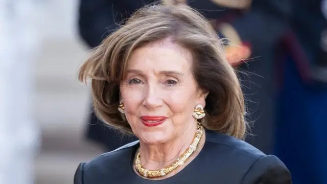 Nancy Pelosi looking at the camera in a dark top with gold jewellery