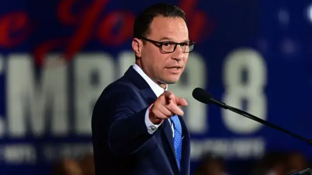 Josh Shapiro, wearing a blue suit, is stood in front of a microphone and pointing towards the camera