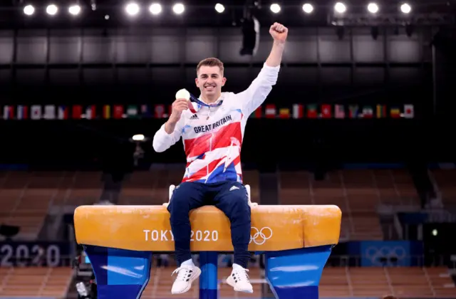 Max Whitlock sits on a pommel horse and holds up a gold medal