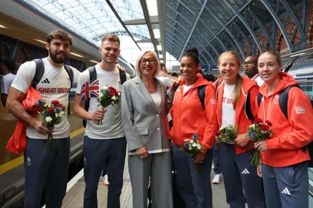 Patrick Brown, Lewis Richardson, Gwendoline Cazenave, Chantelle Reid, Rosie Eccles and Charley Davison stand on a train platform with flowers in their hands