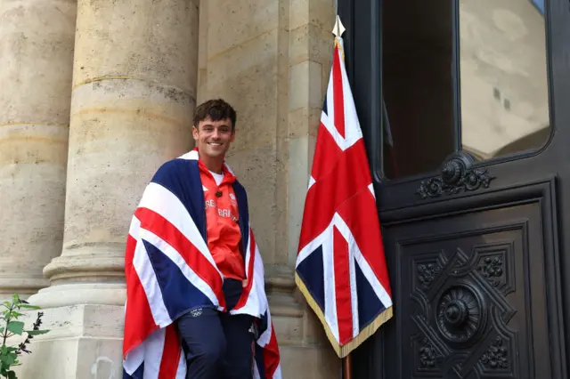 Tom Daley leans against a wall with a Union Jack over his shoulders