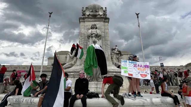 Protestors hold signs and stand around a fountain