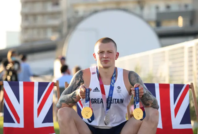 Adam Peaty holds up two gold medals and wears a silver around his neck