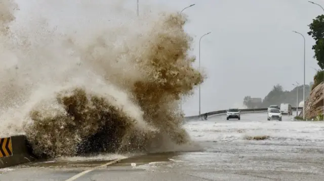 Large wave crashing over a flooded road with cars in the background