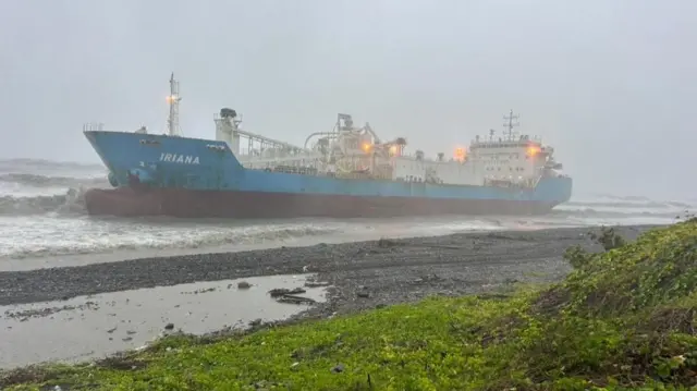 Large cement ship run aground along coast with grassy hill in foreground