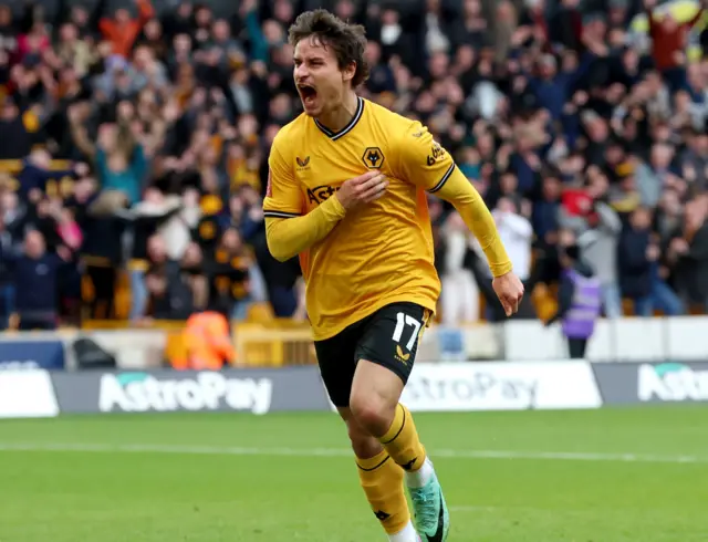 Hugo Bueno runs away in celebration after scoring against Coventry. He is wearing Wolves' Old Gold home shirt and black shorts with green boots. He is putting his right arm to his chest and shouting.