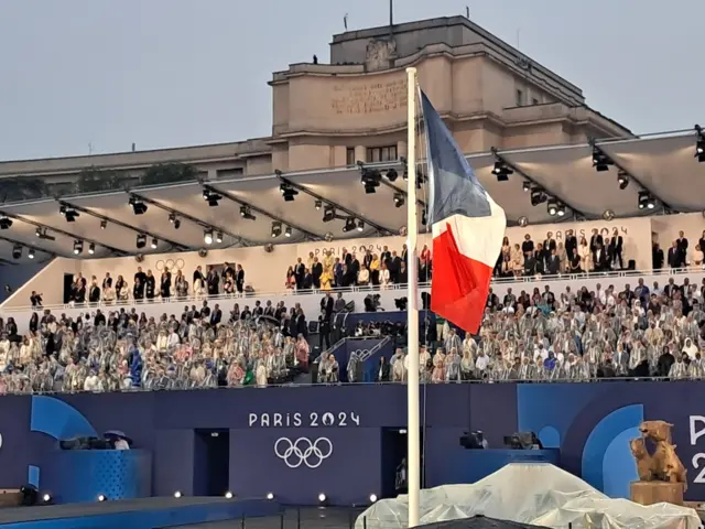 A stand full of spectators at the Paris 2024 opening ceremony. A French flag stands in the foreground.