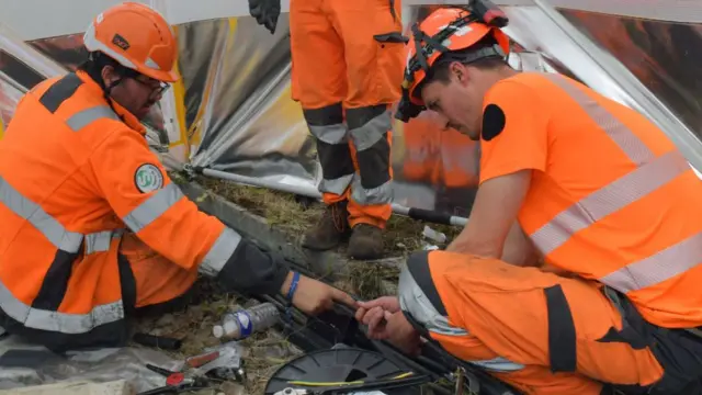 Three rail works wearing hardhats and orange high-vis clothing are working on underground cables