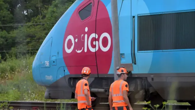 Two workers in orange high-vis clothing watch a blue high-speed train as it moves past them