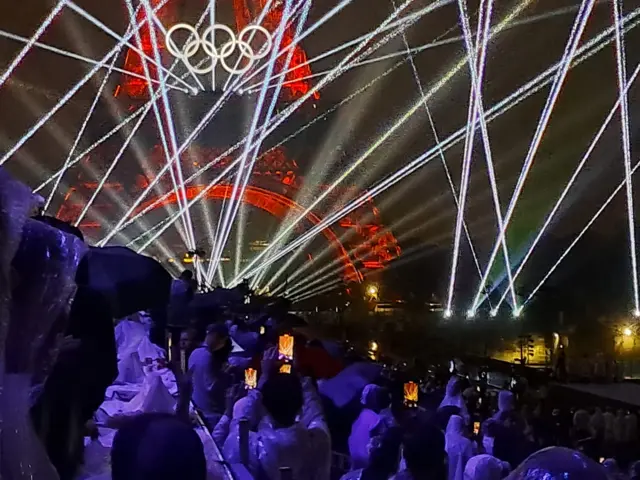 Eiffel Tower bathed in red light and white lasers, as people hold up their smartphones to take pictures.