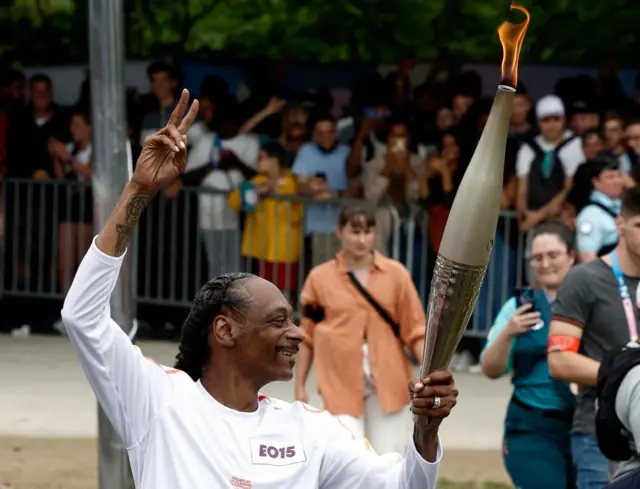 US rapper Snoop Dogg holding the Olympic torch, smiling at crowds and making a peace sign with his other hand