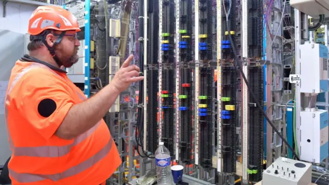 A rail worker in orange high-vis and a hardhat looking at a computer network connections in a server room