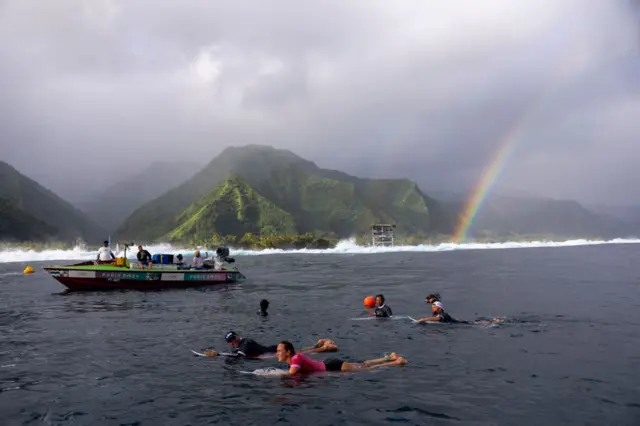 The surfing taking place in Tahiti with mountains and rainbow in the background