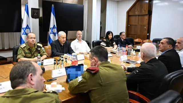 Nine men, wearing military uniforms and suits, and one woman sit at a table with two Israeli flags behind them