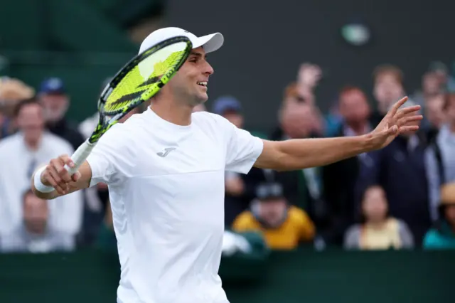 Dressed in all white at Wimbledon, Aleksander Vukic smiles with his arms raised to the side with crowd looking onwards