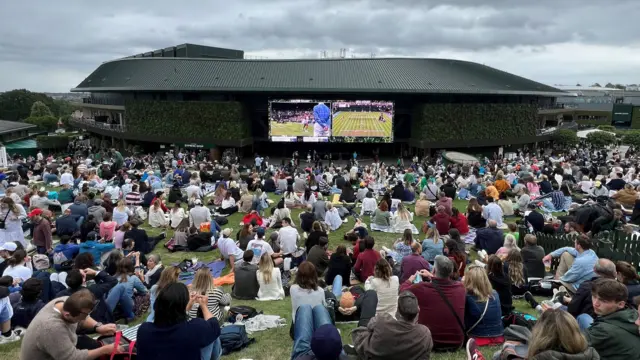 People sitting on a hill watching two tennis matches on a large screen