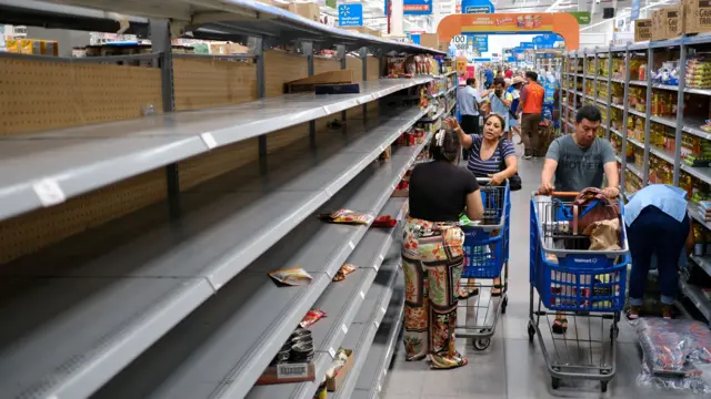 People with shopping trolleys stand in a supermarket aisle with empty shelves