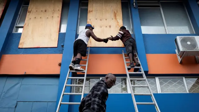 Two workers on ladders board up windows at a blue office building as Hurricane Beryl approaches.