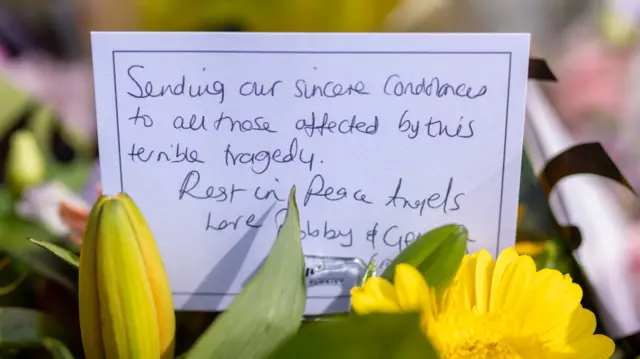 A handwritten note among flowers 'Sending our sincere condolences to all those affected by this terrible tragedy. Rest in Peace Angels.'