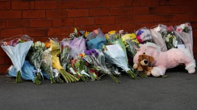 Bouquets of flowers and stuffed animals are placed by a brick wall