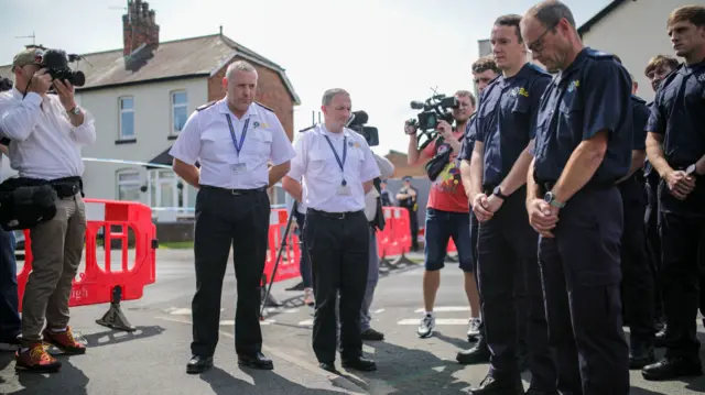 Members of the fire and rescue service stand with heads bowed as they pay tribute at the scene