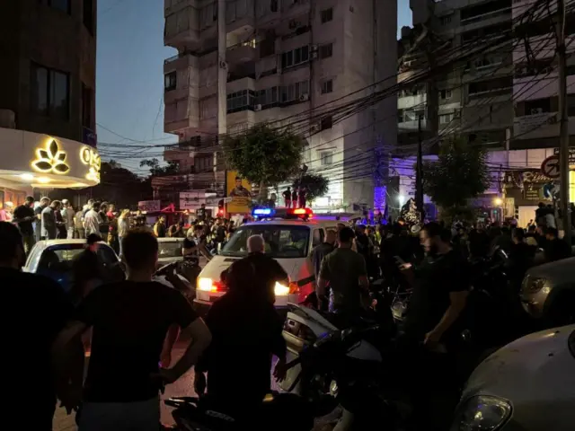 A large crowd of people surround an ambulance on a street, a damaged multi-story building is in the background