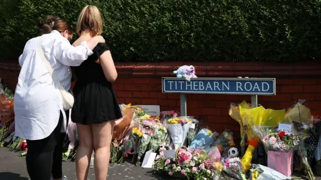 Two women with their backs to the camera look at tributes left at the scene of a stabbing attack