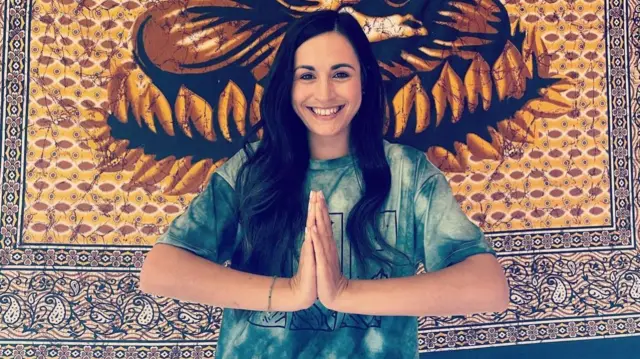 Lucas, a woman with long brown hair, holds her hands in a prayer pose as she smiles at the camera