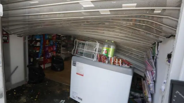 The shutters of the mini mart are bent back - debris and signs of disruption on the floor