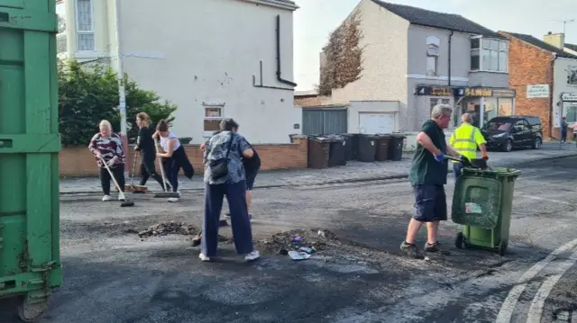 various people sweep a street with brooms