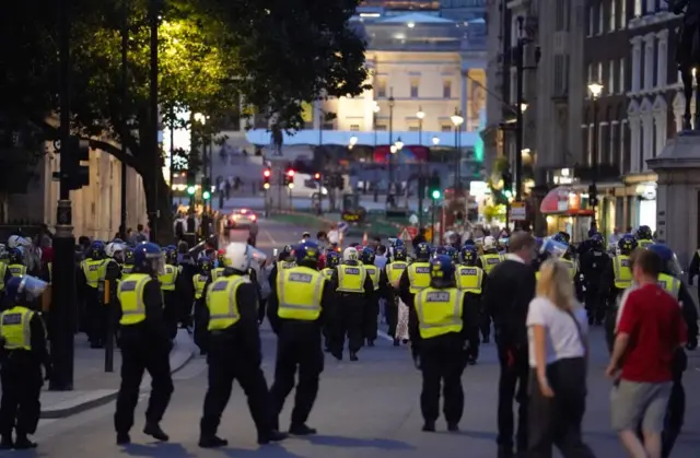 Police in riot gear stand in a road, with protesters behind them