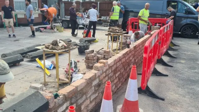 Landscape shot of builders rebuilding the damaged wall. Cones placed in front of wall