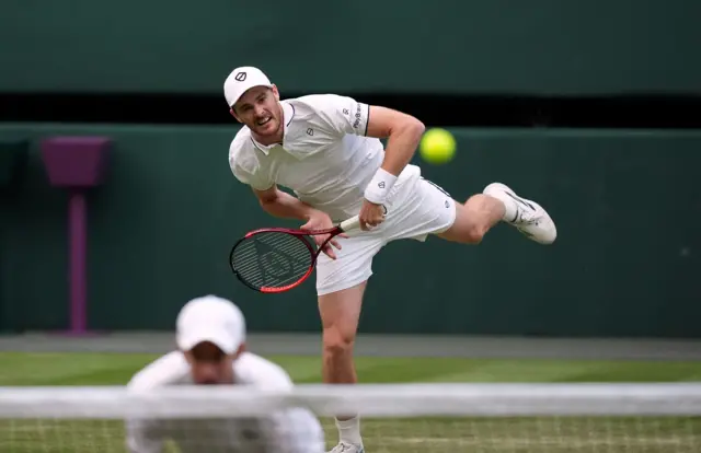 Jamie Murray serves on Centre Court with Andy Murray crouched in front of the net in the foreground, out of focus