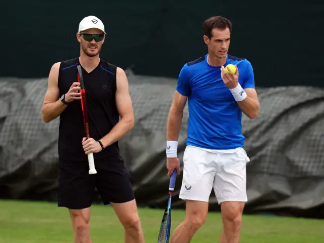 Jamie Murray holds a tennis racquet next to Andy Murray who holds a tennis racquet and a tennis ball