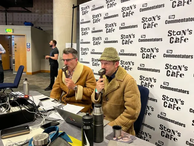 Two men in fleece coats and microphones like a scene from 1970s commentary