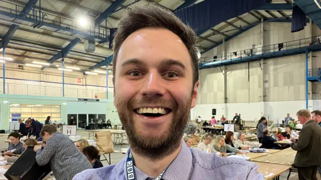 Man smiling in front of lots of people counting votes