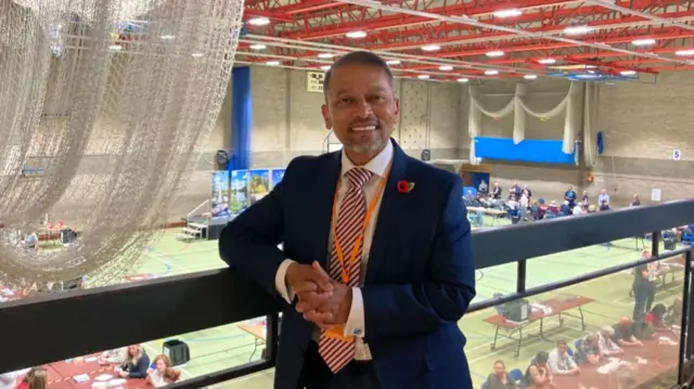 Chan Abraham wearing a suit and stripey tie standing on a balcony with counters in the background.