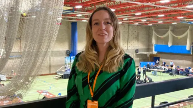 Georgie Hunt standing on a balcony by the count hall wearing a green and black striped jumper and an orange lanyard.