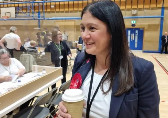 A smiling Lisa Nandy wearing a white top, blue blazer and red rosette. She is in a sports hall and people counting ballots can be seen behind her