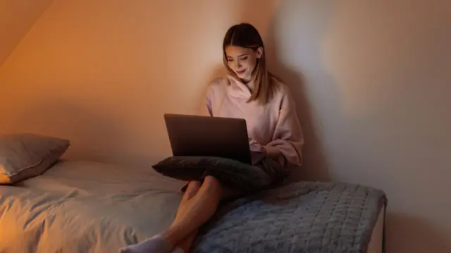 A young woman looks at a laptop on her knee while sitting on a bed