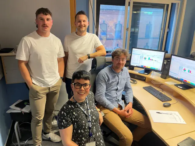 The Results Hub Team stand at a desk in Broadcasting House in Belfast