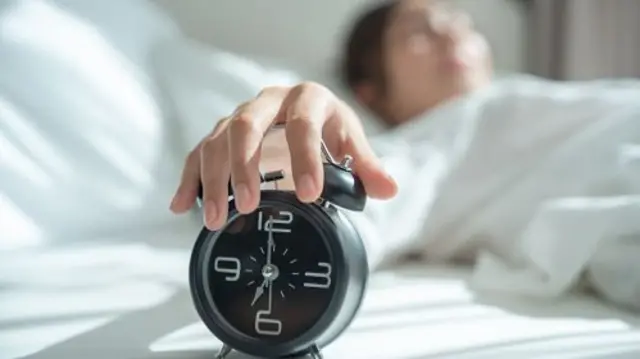 A woman lying in bed reaches out to snooze an alarm clock.