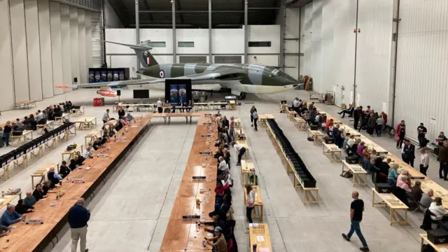 Rows of counters are sat at tables waiting for the ballot boxes to arrive in a hanger, with a large green and blue plane in the background.