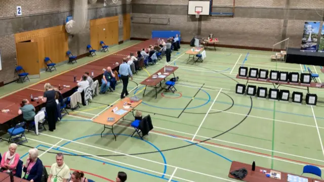 Hall with people sitting at tables and ballot boxes on the right